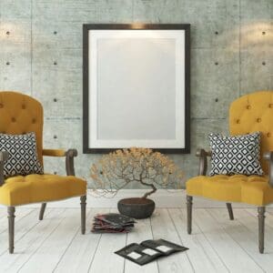 Two yellow armchairs with patterned cushions face each other in a modern room with concrete walls and a large empty framed picture between them.