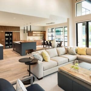 Spacious modern living room with large sofa, open kitchen, dining area, and floor-to-ceiling windows overlooking nature.