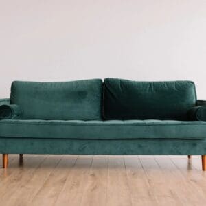 A teal velvet sofa with a minimalist design and wooden legs stands against a plain white wall on a light wooden floor.