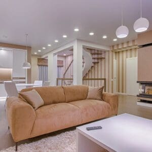 Modern living room with a beige sofa, white coffee table, and a spiral staircase in a well-lit apartment with kitchen visible in the background.