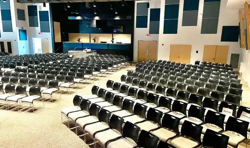 Empty auditorium with rows of metal chairs facing a stage and podium, featuring blue wall accents and acoustic panels.