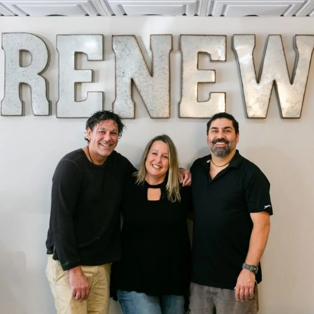 Three smiling adults, two men and a woman, stand in front of a wall with the word "renew" in large metal letters.